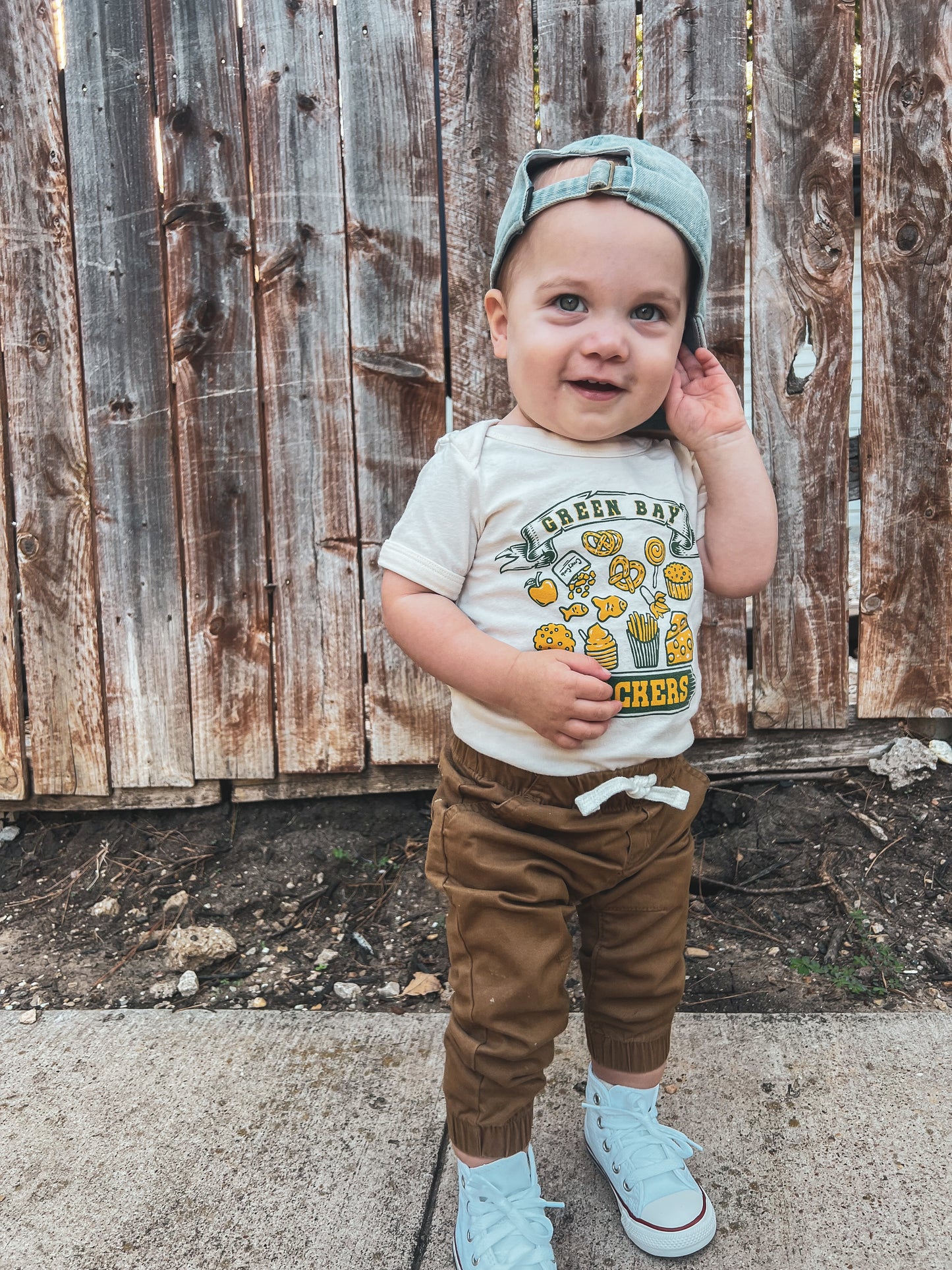 toddler wearing a green bay snackers onesie standing in front of a fence