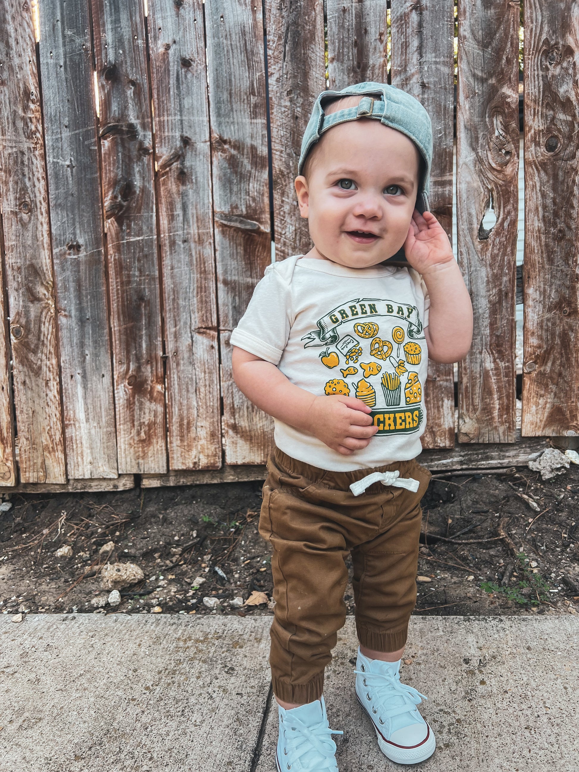 toddler wearing a green bay snackers onesie standing in front of a fence